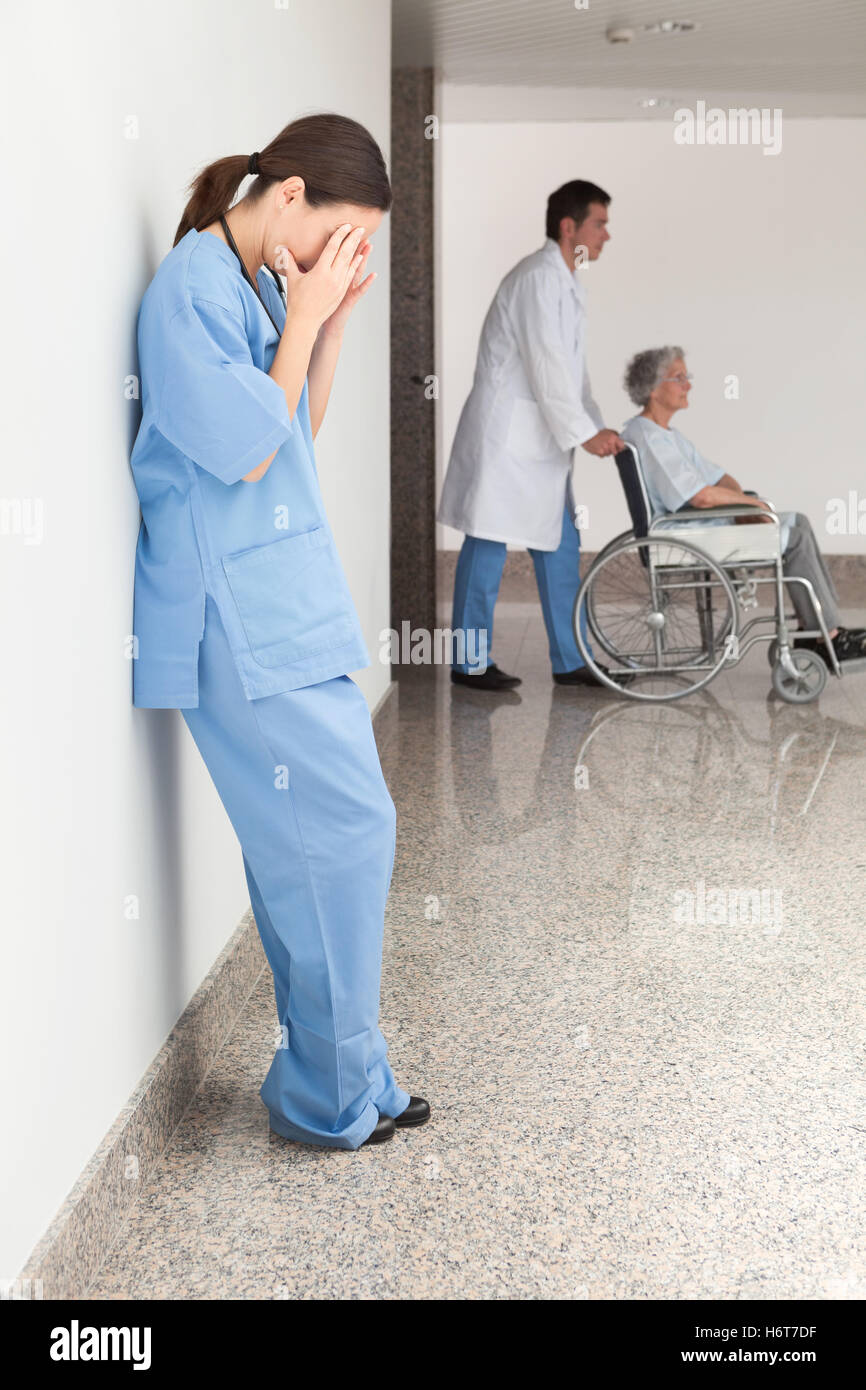 Stressed nurse leaning against wall with doctor pushing patient in wheelchair Stock Photo