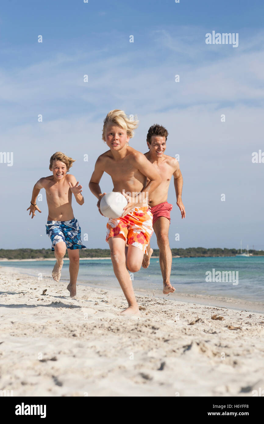 Boy running with rugby ball chased by brother and father on beach, Majorca, Spain Stock Photo