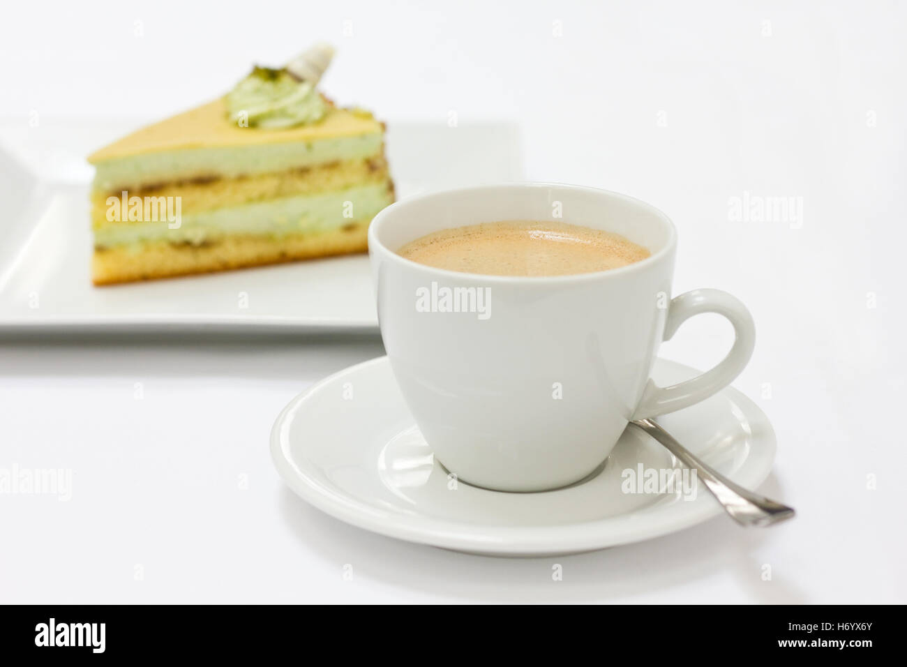 Coffee in a white cup with a spoon and a slice of cake slightly blurry in the background Stock Photo