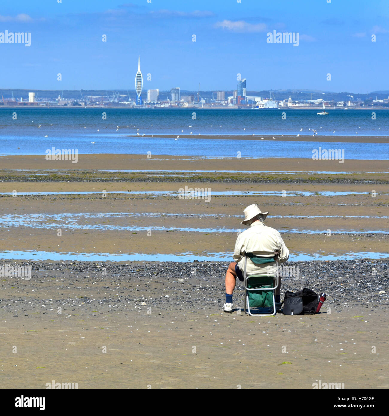 Older man sitting alone on Ryde Isle of Wight UK beach at low tide sketching views of the Solent towards distant Portsmouth on hot summer blue sky day Stock Photo