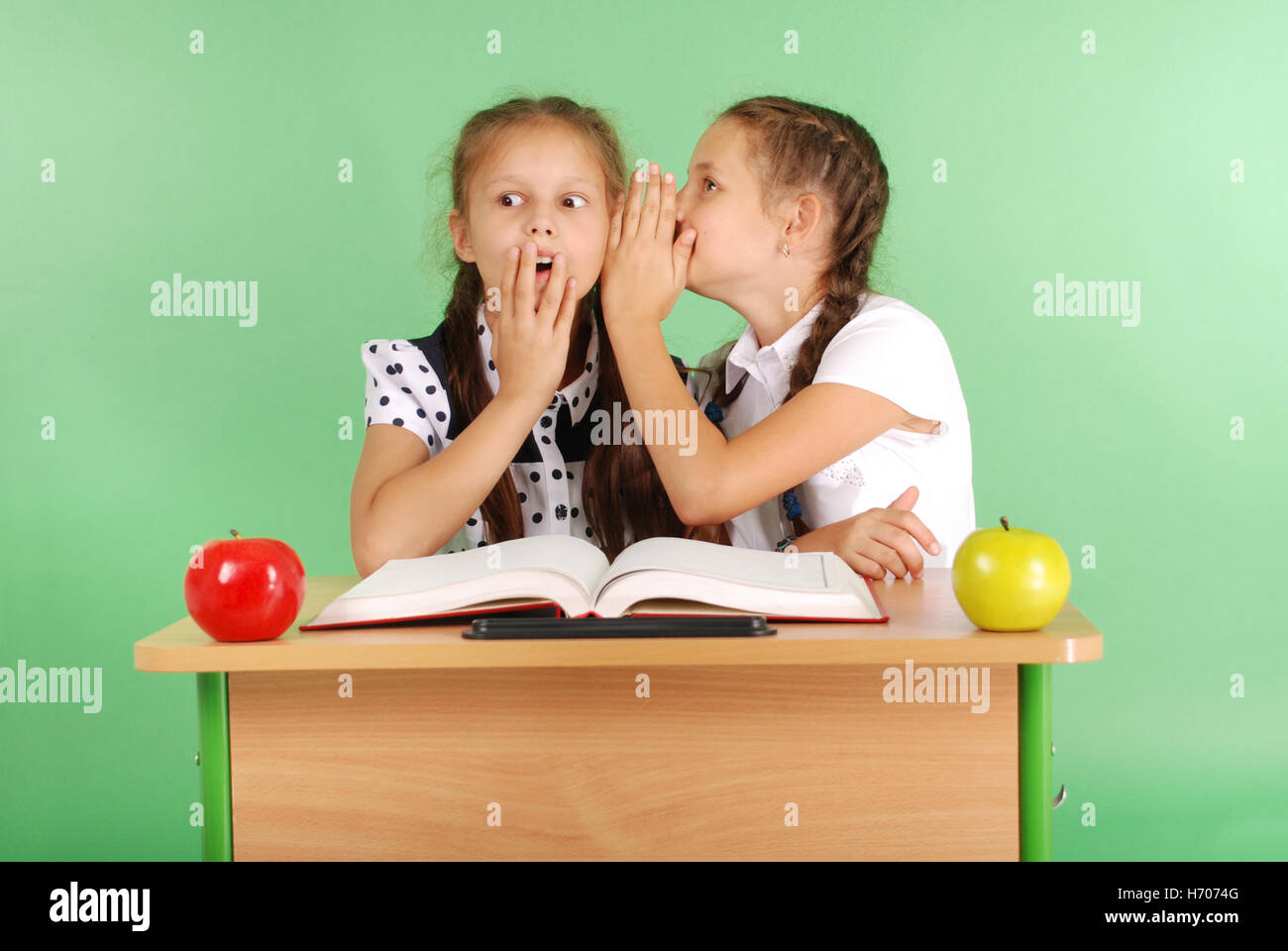 Two school girl sharing secrets  sitting at a desk from book isolated on green Stock Photo