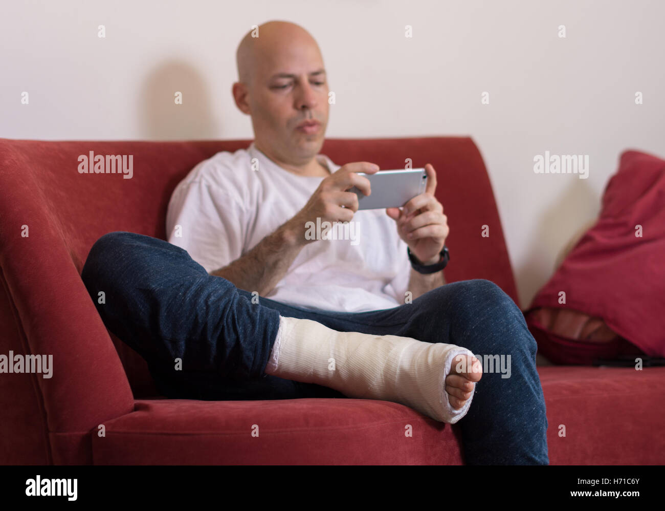 Young man with a broken ankle and a white cast on his leg, sitting on a red couch, surfing the web on his phone (selective focus Stock Photo