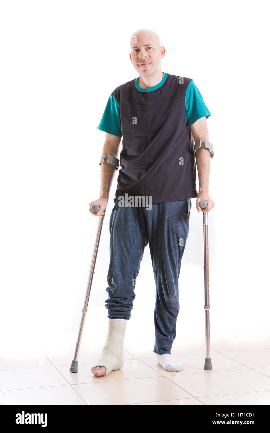 Young man with a broken ankle and a fiberglass and plaster cast on his leg, leaning on his crutches (isolated on white, full bod Stock Photo