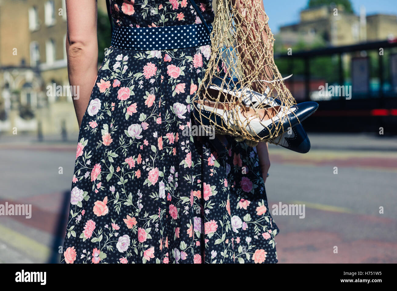 A young woman wearing a dress is standing in the street with a net bag containing a pair of spare party shoes Stock Photo