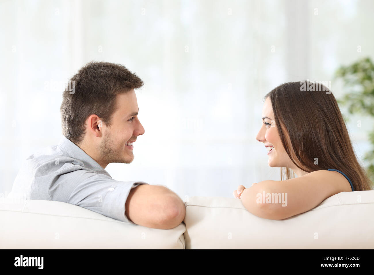Side view of a couple or marriage talking sitting on a couch and looking each other at home with a white curtains Stock Photo
