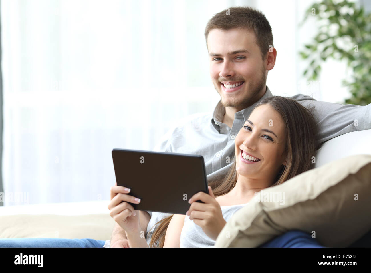 Couple holding a tablet posing sitting on a couch at home and looking at camera Stock Photo