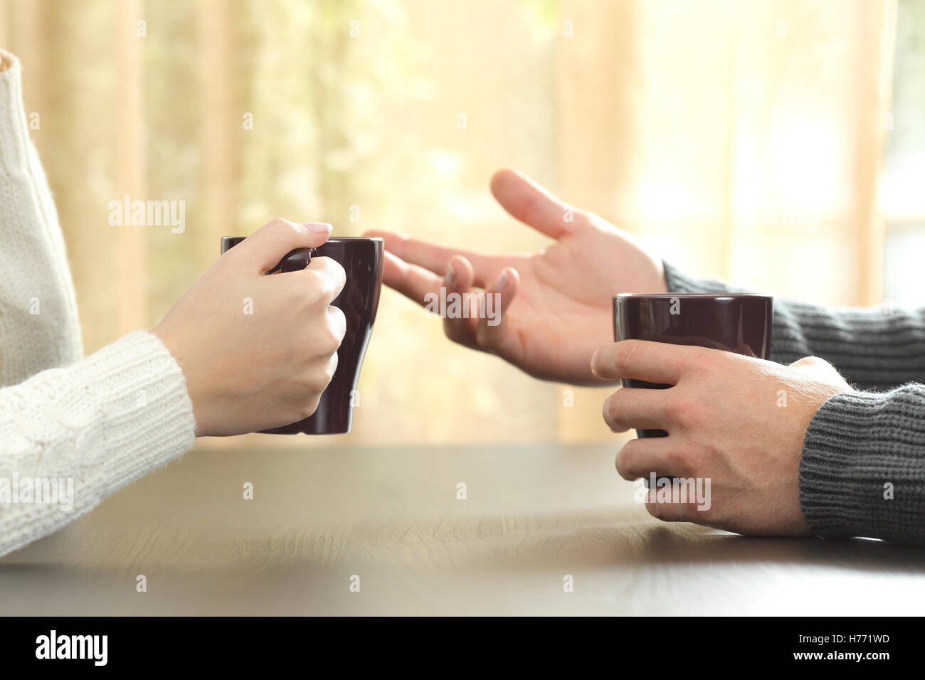 Back light profile of hands of 2 friends or couple talking holding coffee cups sitting in a table at home Stock Photo