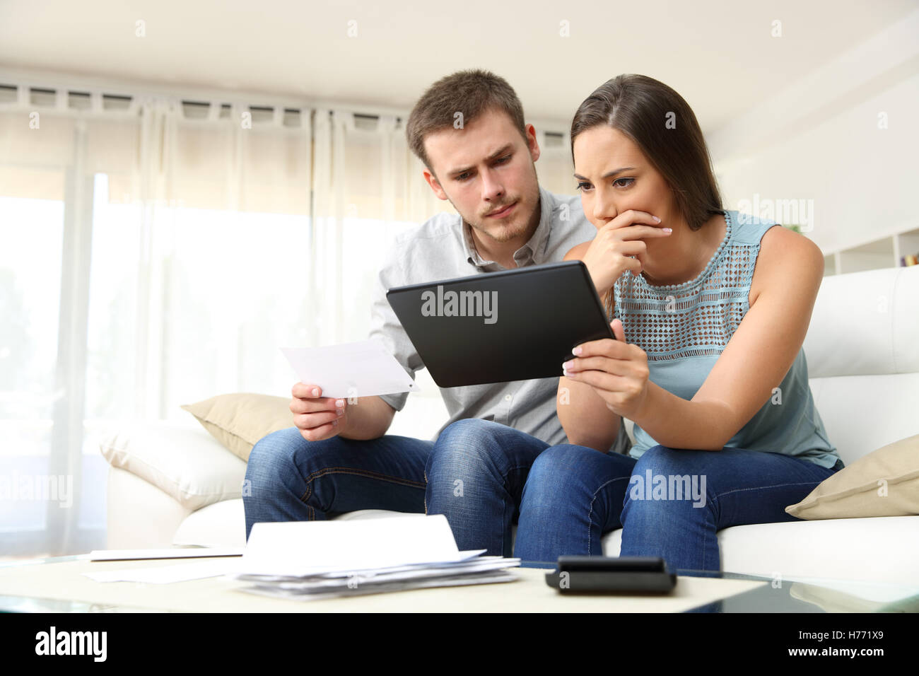 Worried couple checking bank account trouble online in a tablet sitting on a couch in the living room at home Stock Photo