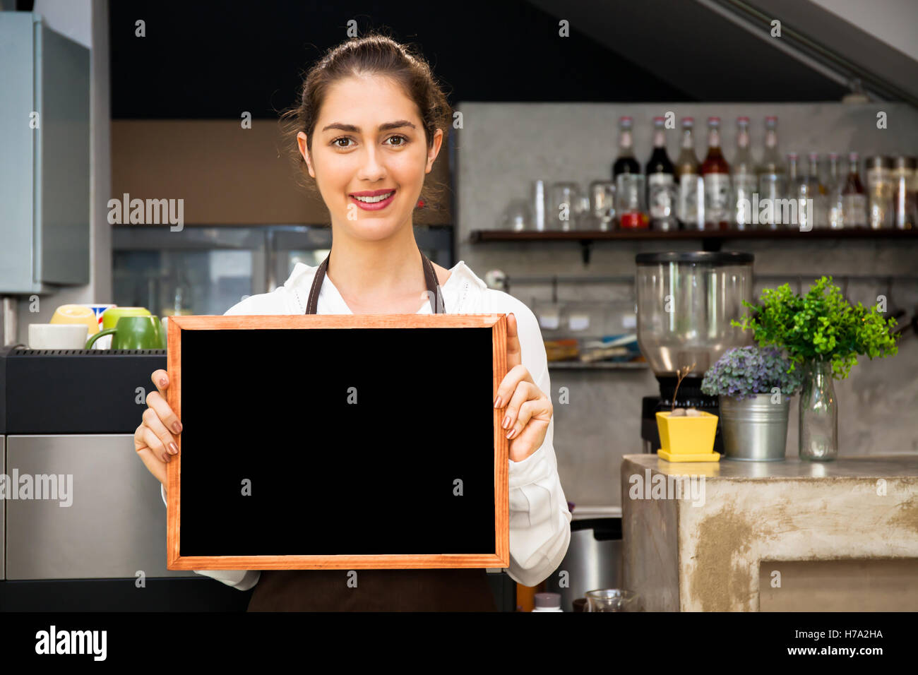Beautiful Caucasian woman in barista apron holding empty blackboard sign inside coffee shop - ready to insert text Stock Photo