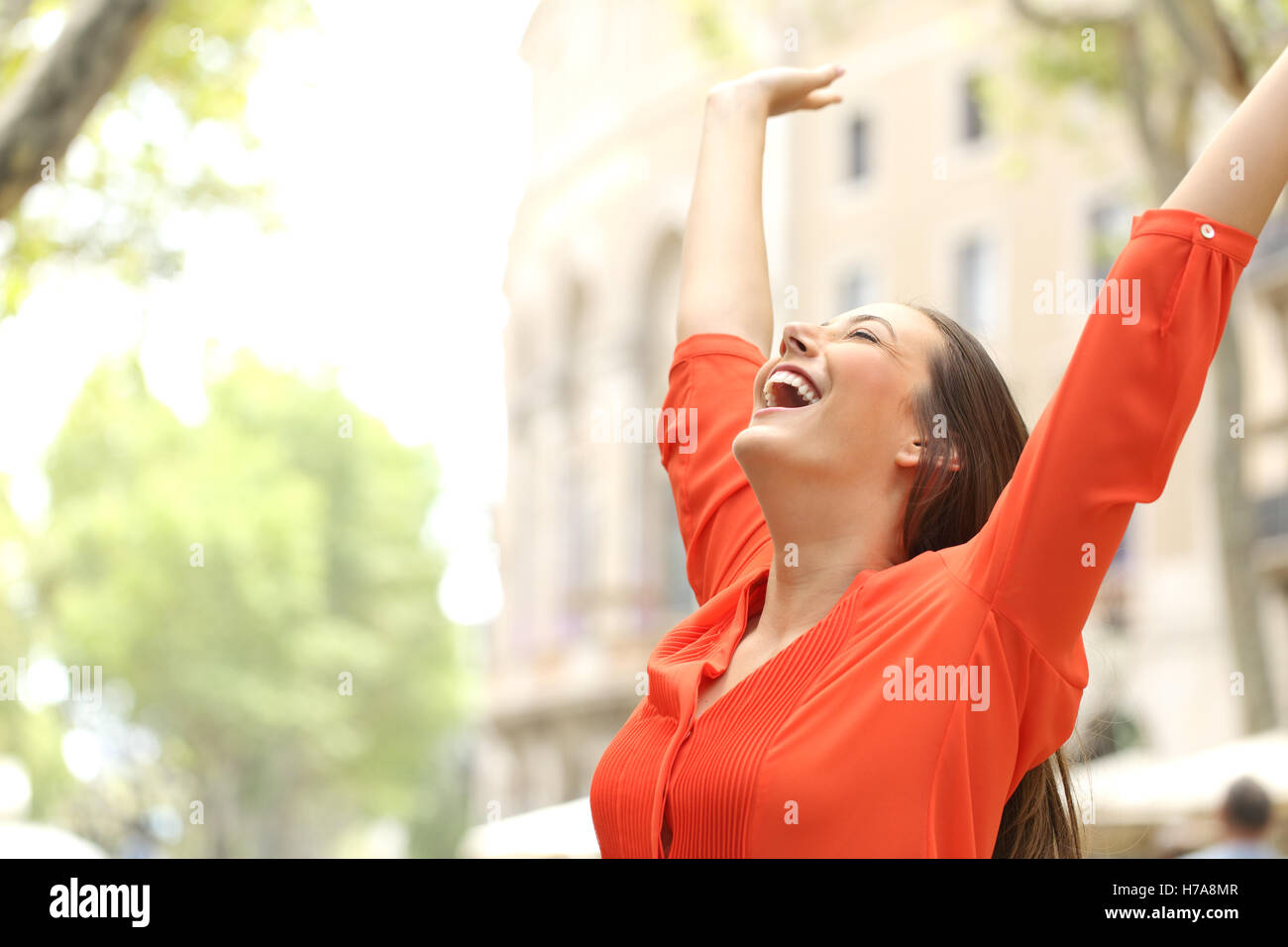 Excited woman wearing orange blouse raising arms outdoors in the street ...