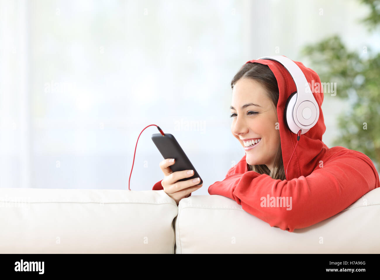 Portrait of a happy teen listening music on line from smart phone sitting in a couch at home with white curtains Stock Photo