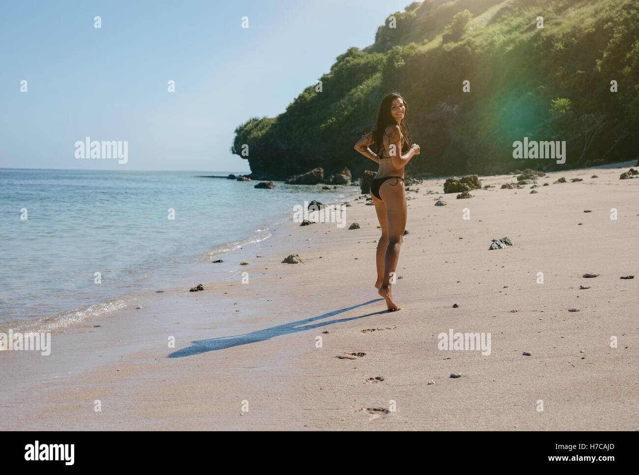 Full length shot of young woman in bikini running on the sea shore and looking back. Beautiful female model enjoying holidays on Stock Photo