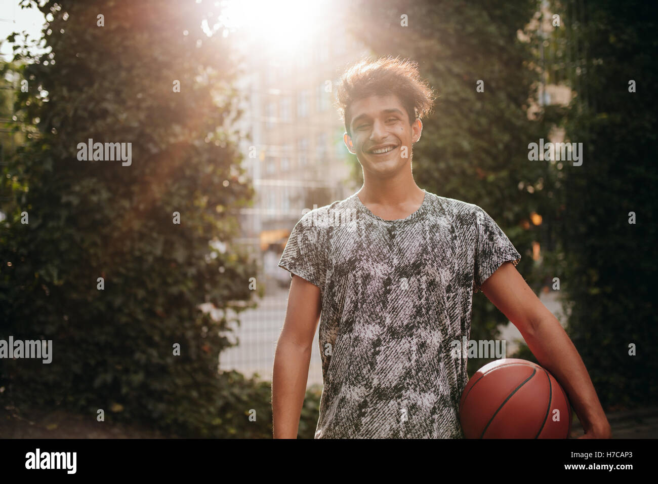 Portrait of handsome young basketball player holding a ball on outdoor court. Smiling teenage streetball player looking at camer Stock Photo