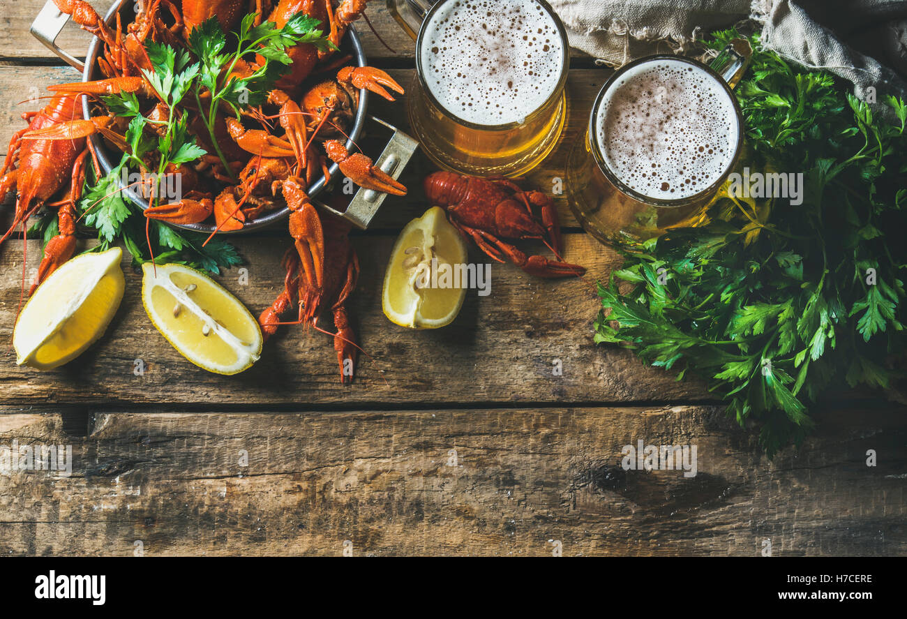 Two mugs of wheat beer and boiled crayfish in pan served with with lemon and parsley over rustic wooden background, top view, co Stock Photo