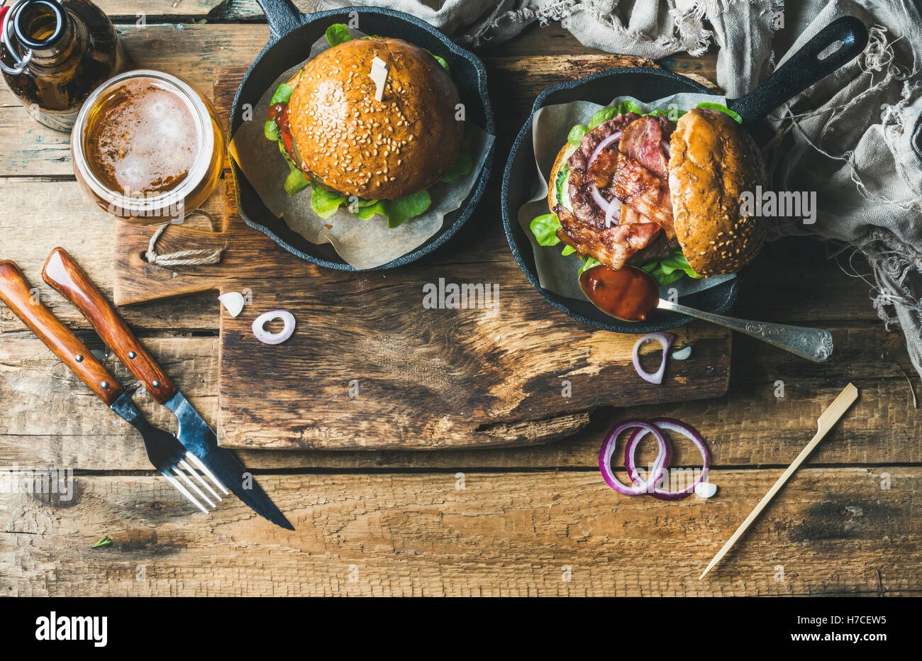 Homemade beef burgers with crispy bacon and vegetables in small pans and glass of wheat beer on rustic serving board over shabby Stock Photo
