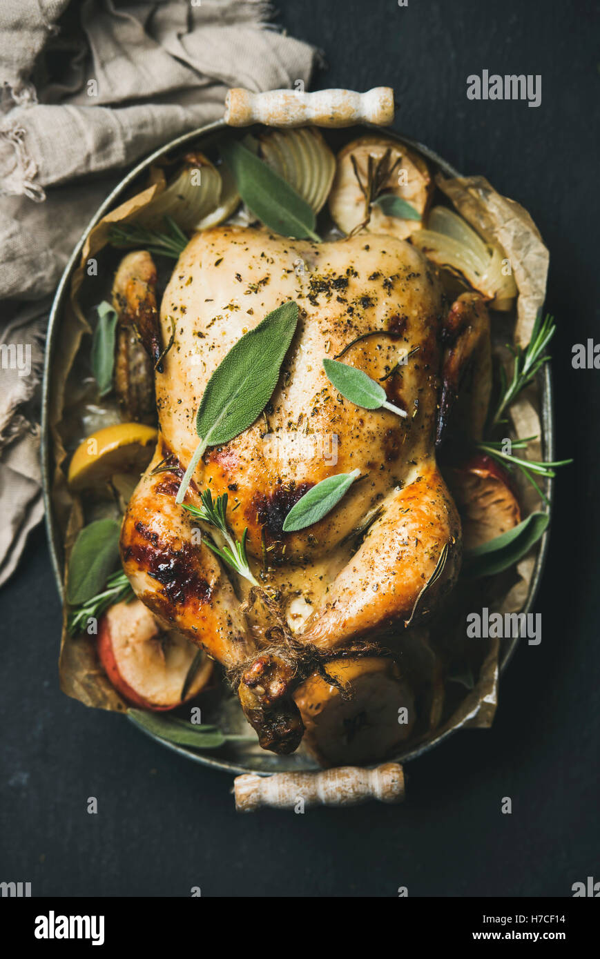 Oven roasted whole chicken with onion, apples and sage leaves in serving tray over dark stone background, top view, selective fo Stock Photo