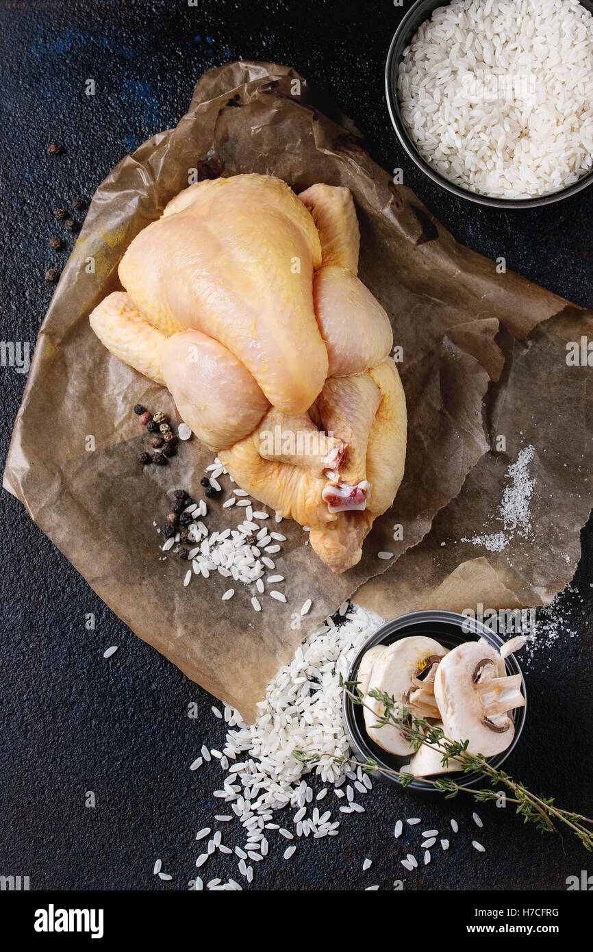 Whole raw mini chicken on baking paper with seasoning, thyme, sliced mushrooms and bowl of uncooked white rice over black textur Stock Photo