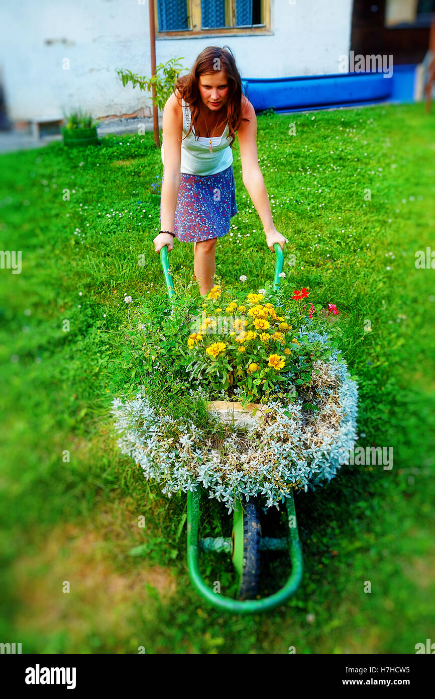 young woman in garden playfully pushing decorative wheelbarrow full of ...