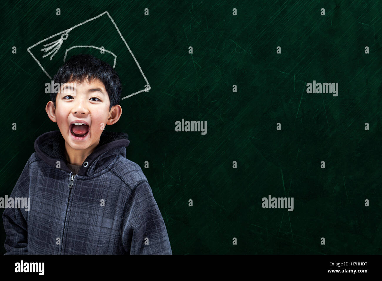 Jubilent Asian boy in classroom setting with graduate hat on chalkboard background and copy space. Concept for university educat Stock Photo