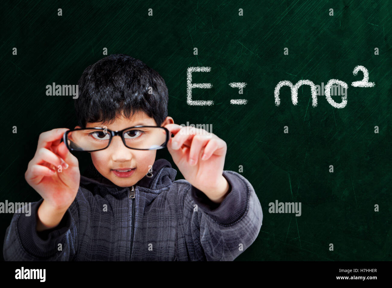 Smart Asian boy holding up eyeglasses on chalkboard background with mathematical equation and copy space. Stock Photo