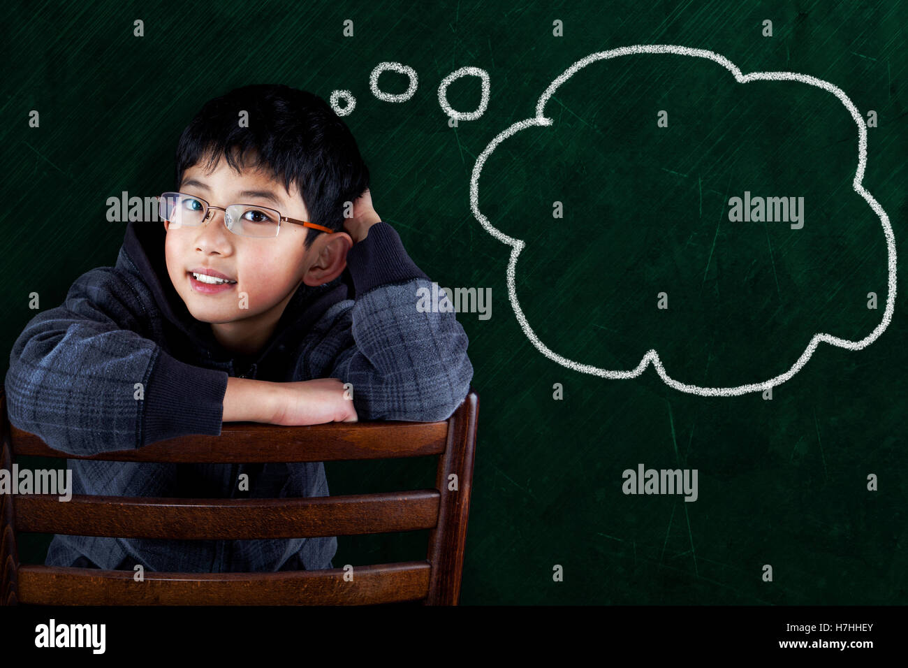 Smart Asian boy sitting on classroom chair with chalkboard background and thought bubble copy space. Stock Photo