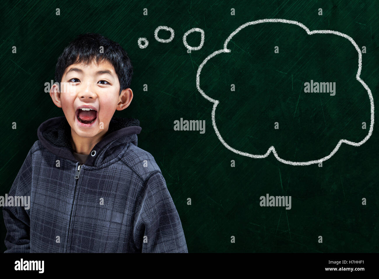Smart Asian boy mouth wide opened in classroom setting with chalkboard background and thought bubble copy space. Stock Photo