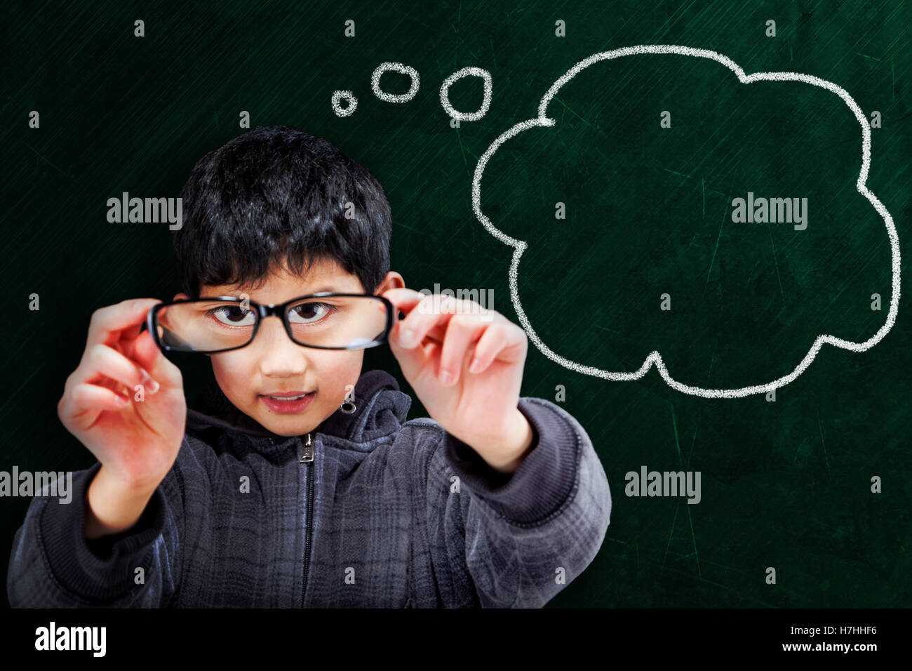 Smart Asian boy holding up eyeglasses on chalkboard background with thought bubble and copy space. Stock Photo