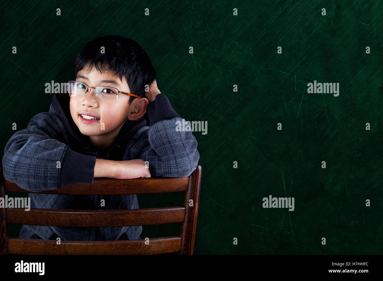 Smart Asian boy sitting on classroom chair with chalkboard background and copy space. Stock Photo