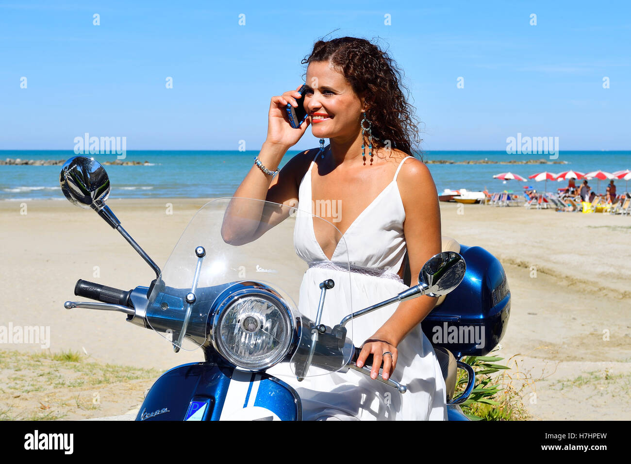 Woman in white dress and decorative earrings with cell phone on Vespa Primavera scooter on the beach, Senigallia, Adriatic Coast Stock Photo