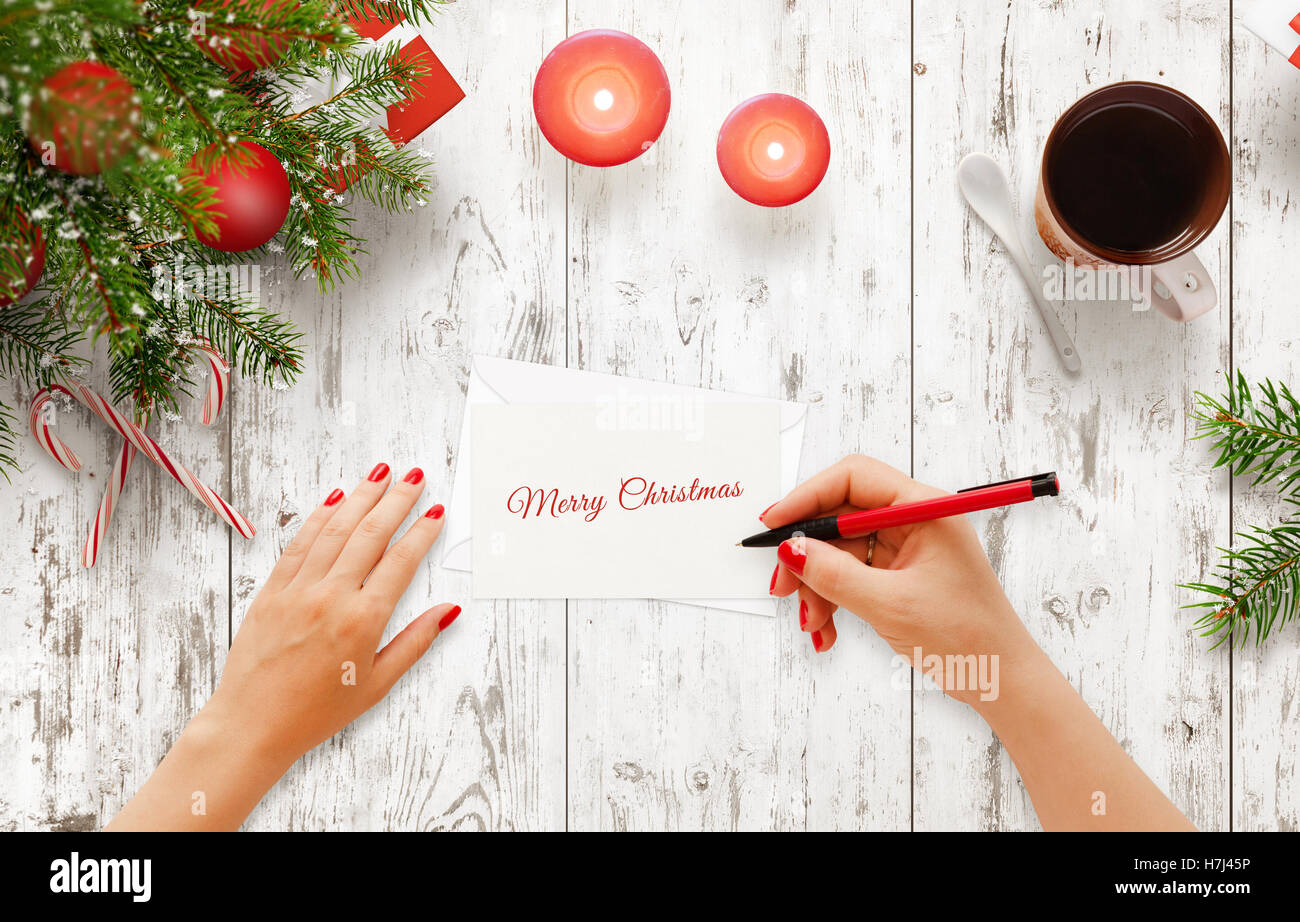 Woman write Christmas greeting message on paper. Top view of white wooden table with christmas tree, decorations, candle, tea Stock Photo