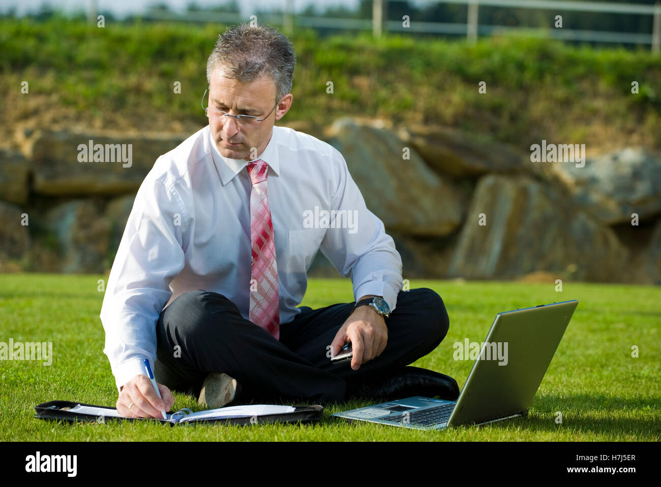 Businessman sitting in a park while using a laptop Stock Photo - Alamy