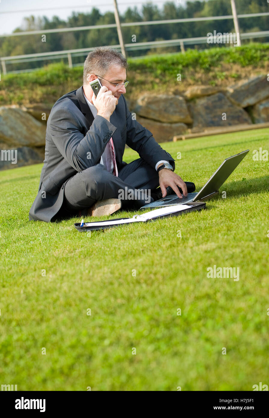 Businessman sitting in a park while using a laptop Stock Photo - Alamy