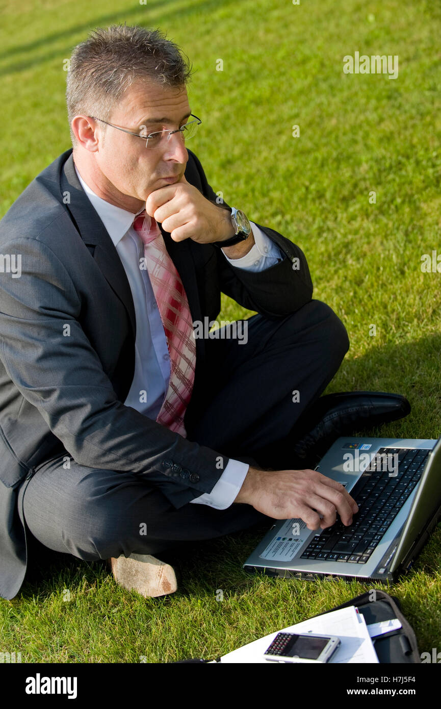 Businessman sitting in a park while using a laptop Stock Photo - Alamy