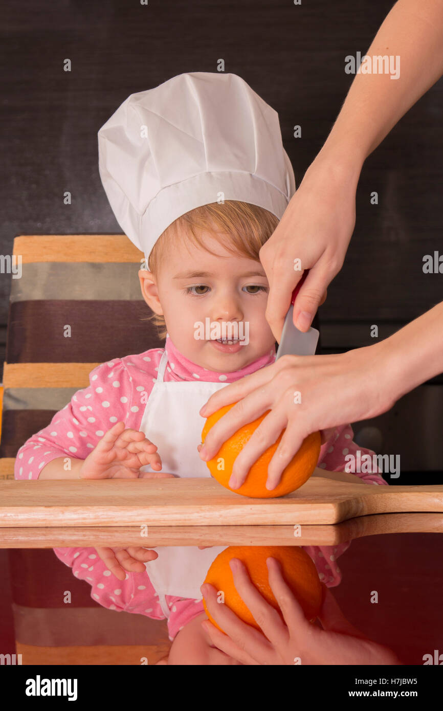 little cute kid in a cook cap playing in the kitchen. Mom cut orange