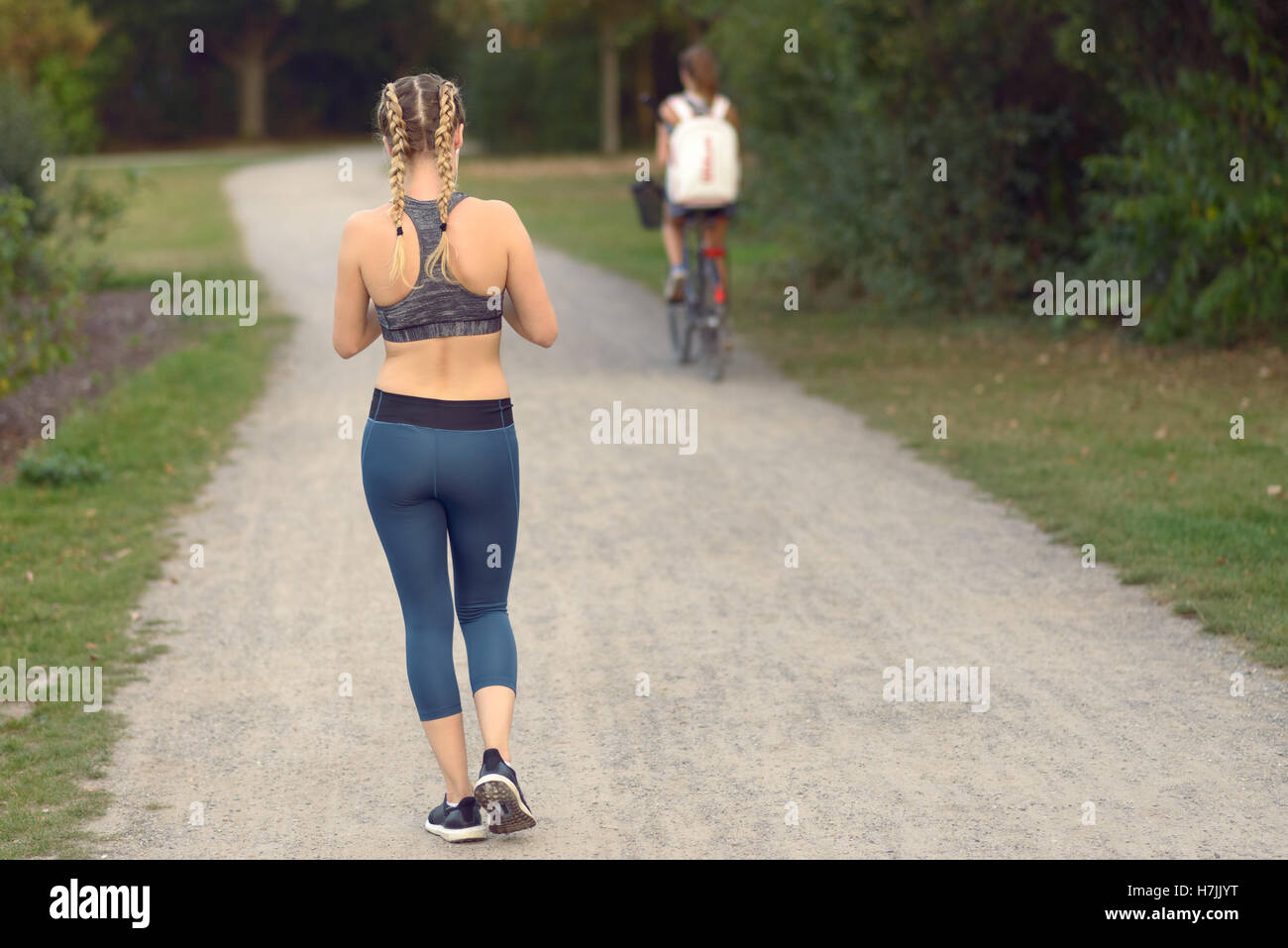 Young woman out jogging in a path moving away from the camera along a narrow lane with a cyclist ahead of her in a healthy lifes Stock Photo