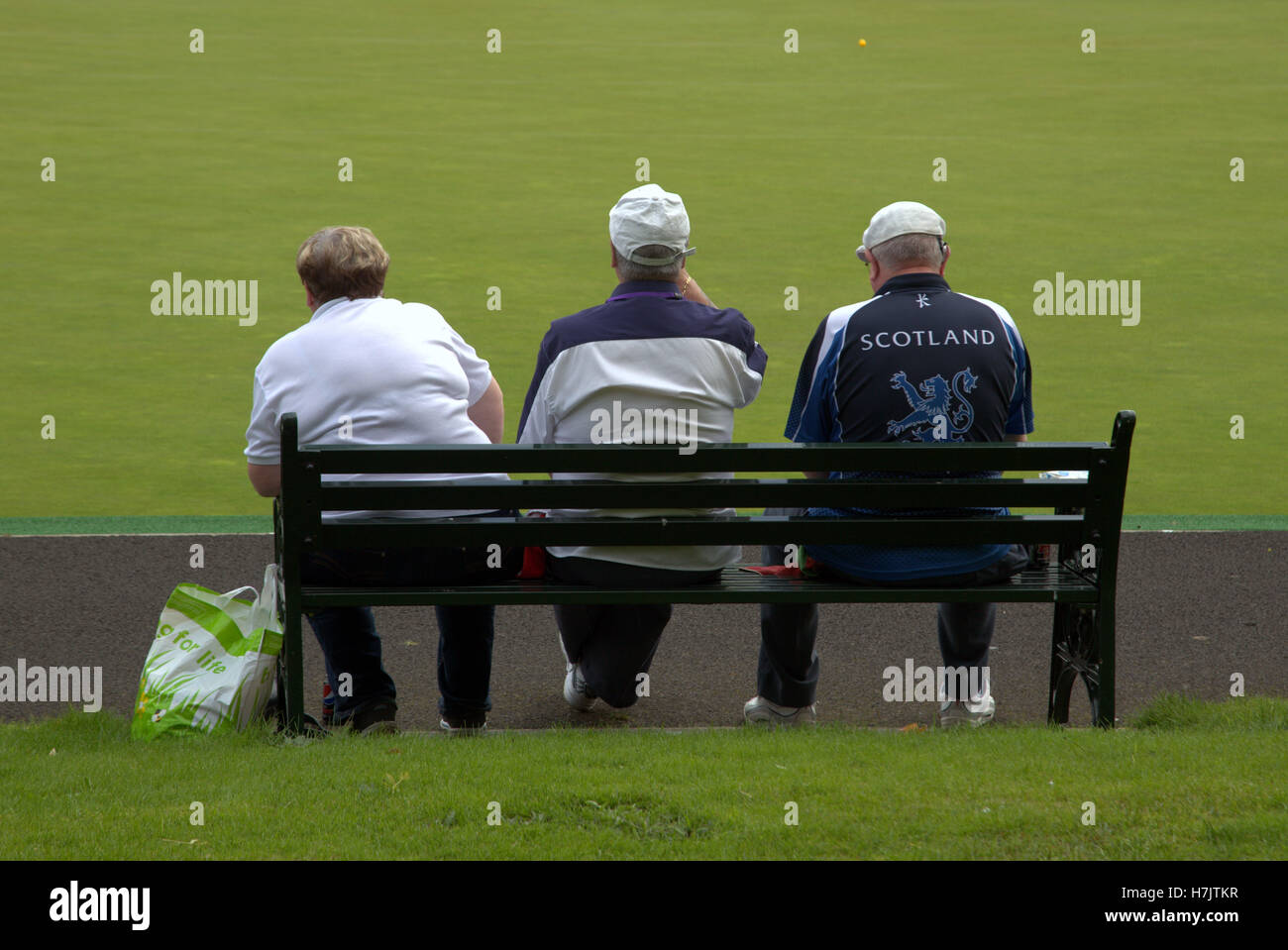three men sitting on a bench with large Scotland logo jacket lawn background Stock Photo