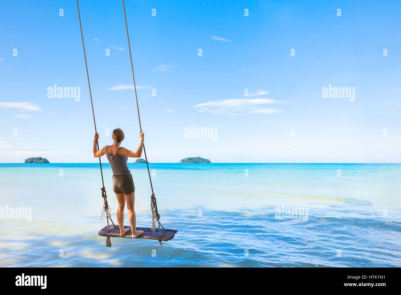 Happy girl swinging and relaxing on a rope swing with a paradise tropical sea in background - concept about summer vacations Stock Photo