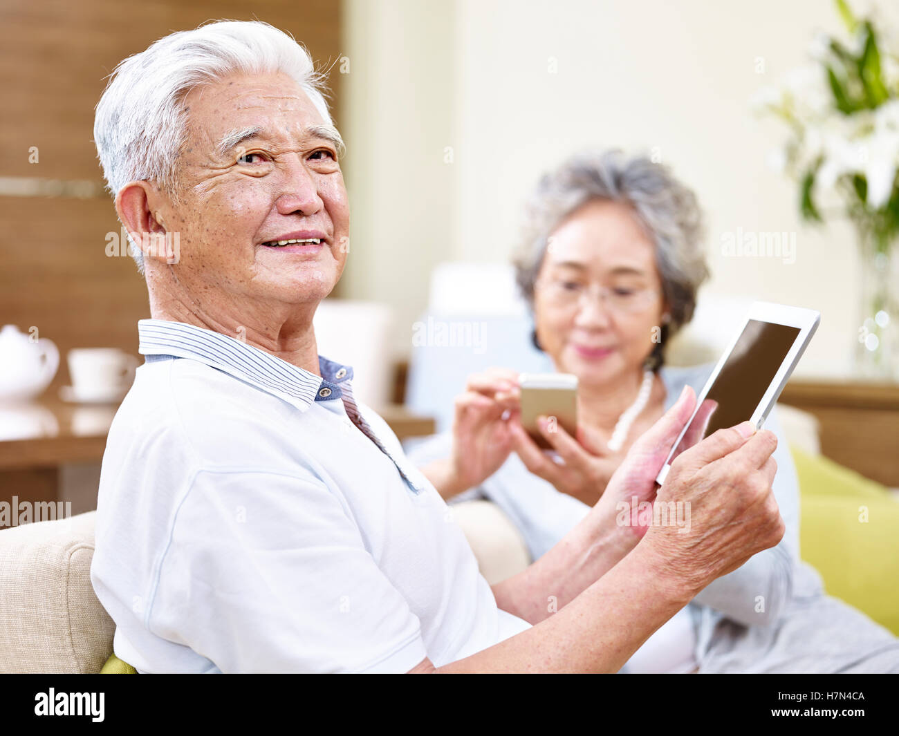 senior asian couple sitting on couch enjoying modern technology using tablet computer and cellphone Stock Photo