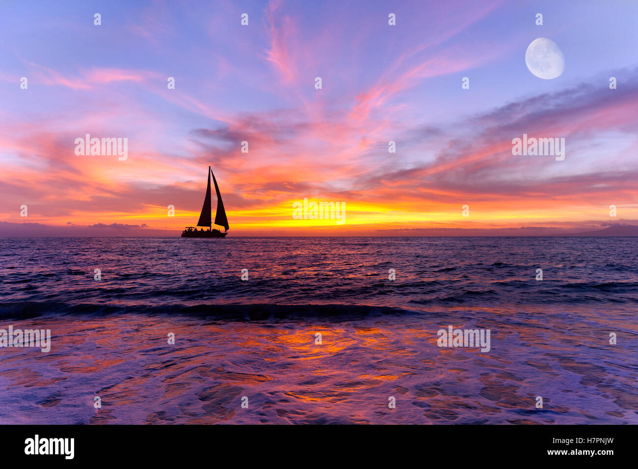 Sailboat ocean sunset is a silhouette of a sailboay sailing along the ocean water with a colorful vivid sunset sky and the moon Stock Photo