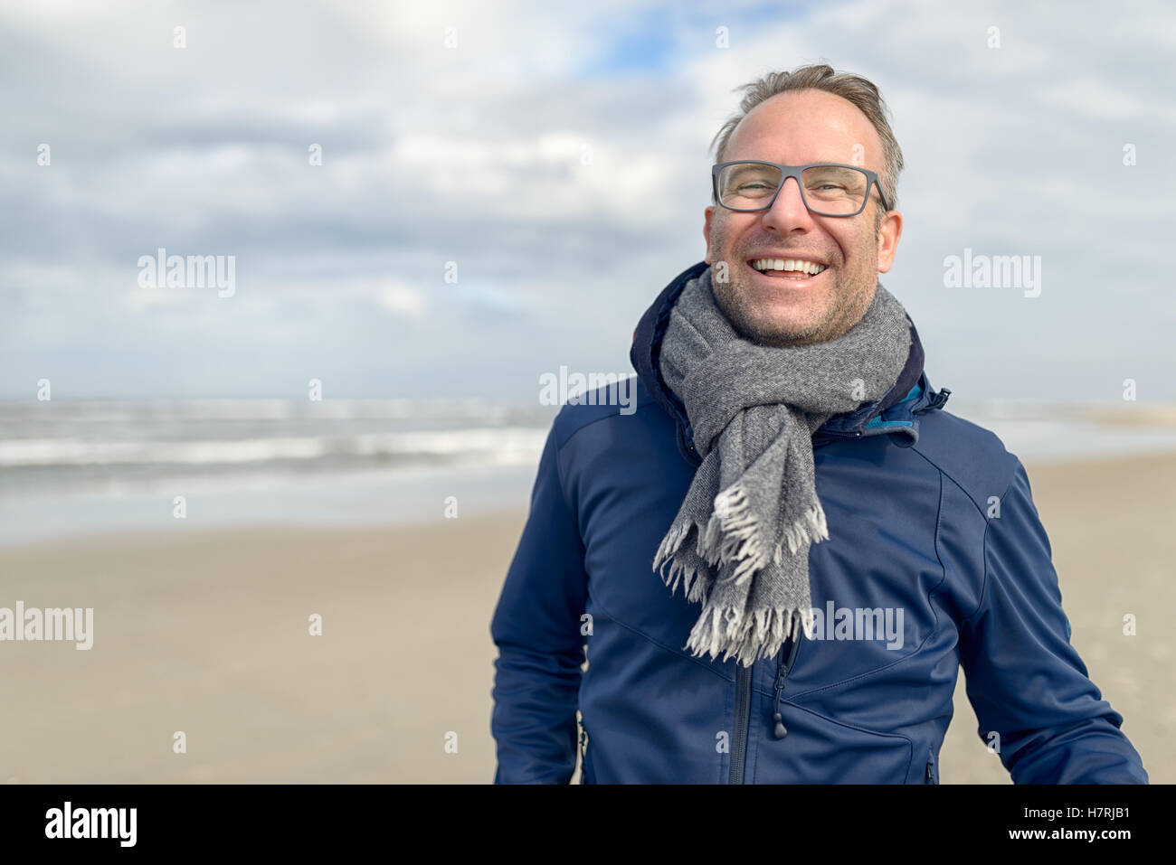 Happy laughing middle-aged man wearing glasses and a knitted woollen scarf standing on a deserted autumn beach on a cloudy day w Stock Photo