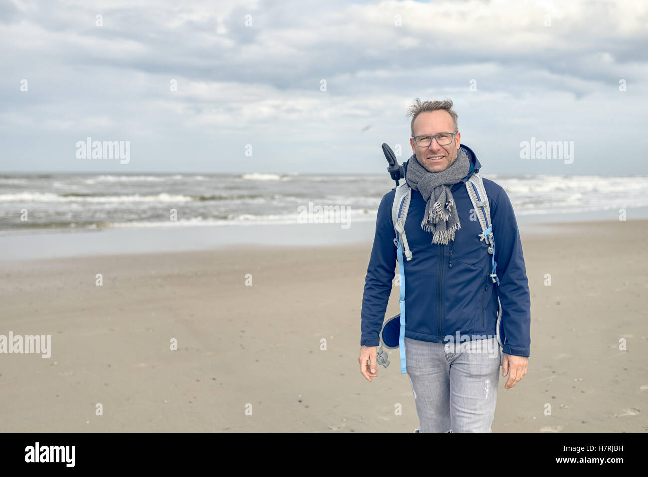 Middle-aged man wearing glasses, a backpack and a scarf walking along a cold blustery beach on a rainy overcast autumn or winter Stock Photo
