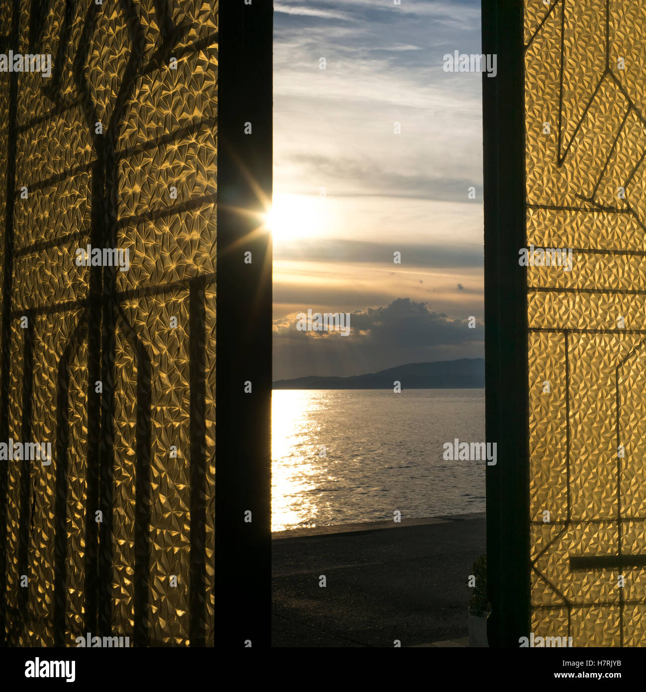 Sunlight glowing at sunset and reflected in the tranquil ocean water as viewed through a window with gold and black shutters Stock Photo