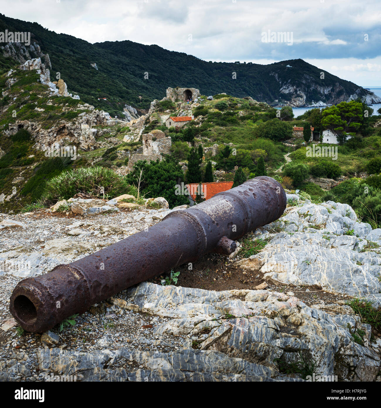 An old, rusted relic laying on the rock on a greek island with a rugged landscape and remote houses in the background; Skiathos, Greece Stock Photo