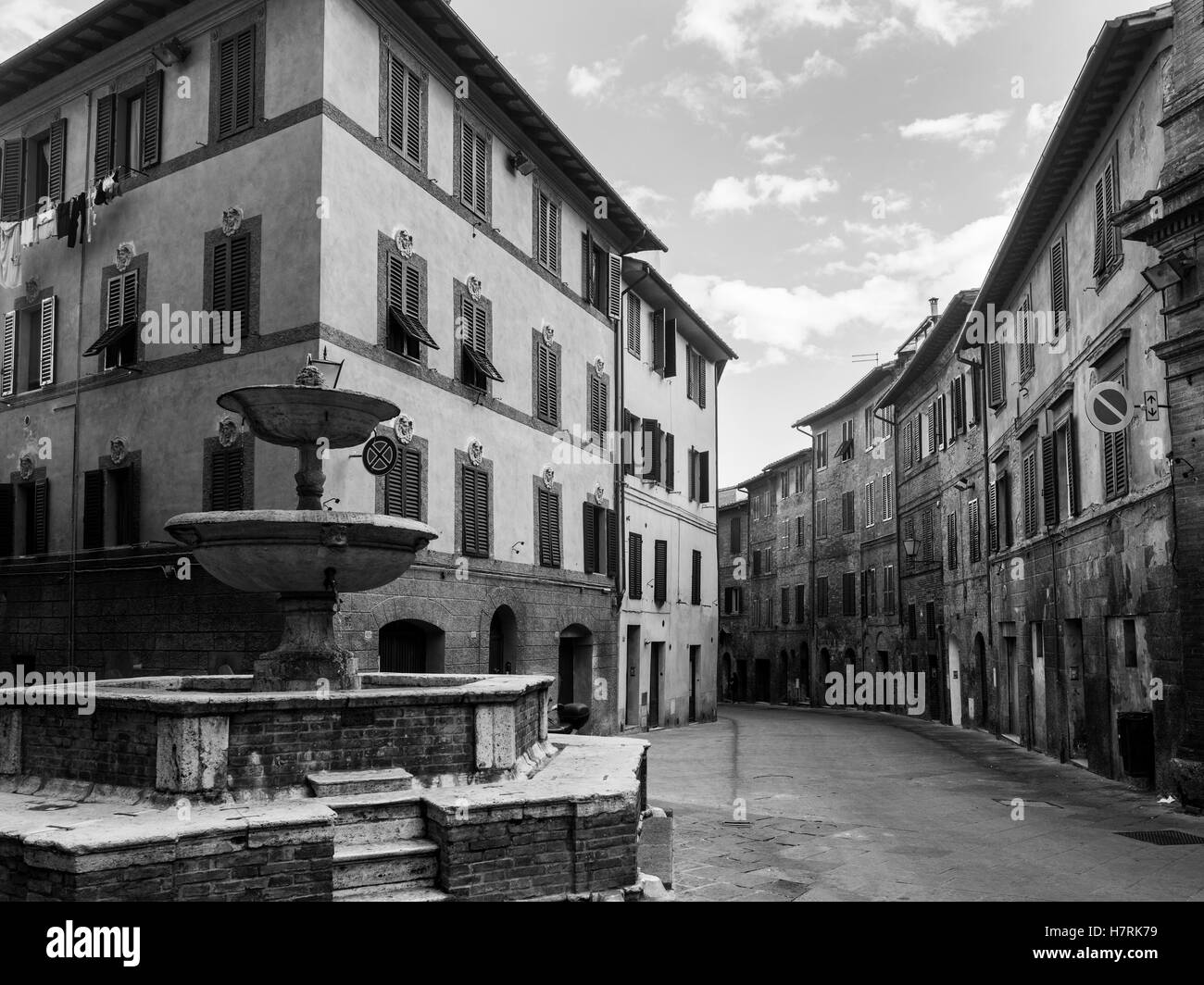 An empty street between two residential buildings and a water fountain in the foreground; Siena, Italy Stock Photo
