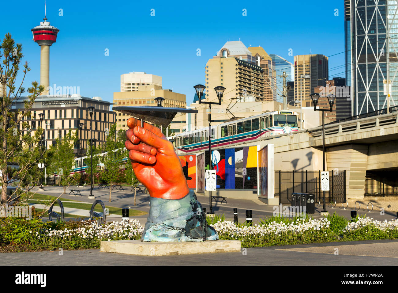 Large hand sculpture holding a metal (paper) plane, with commuter train, city buildings and the Calgary Tower in background with blue sky Stock Photo