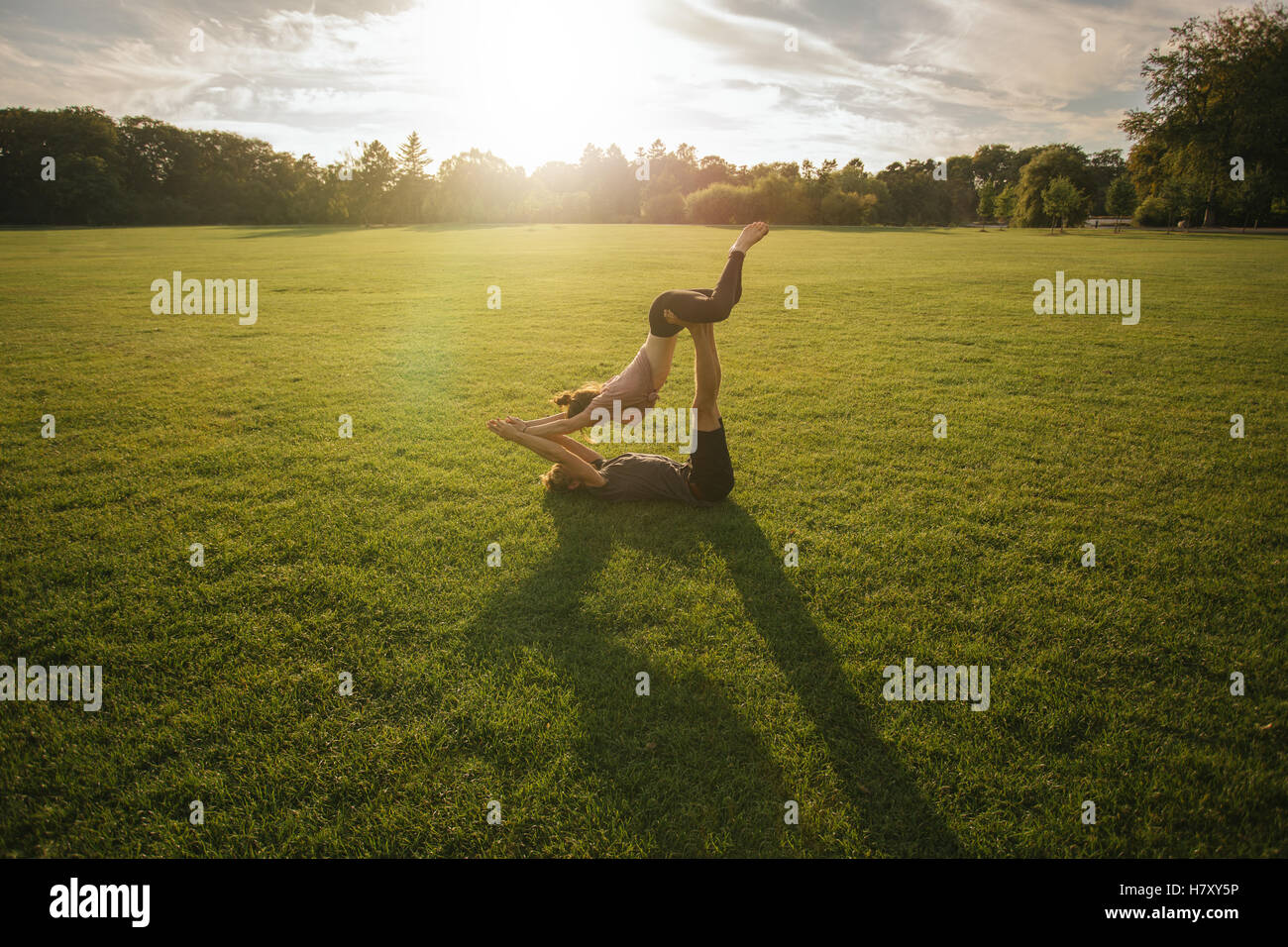 Outdoor shot of young man and woman doing yoga in pair. Couple doing acrobatic yoga workout on lawn. Stock Photo
