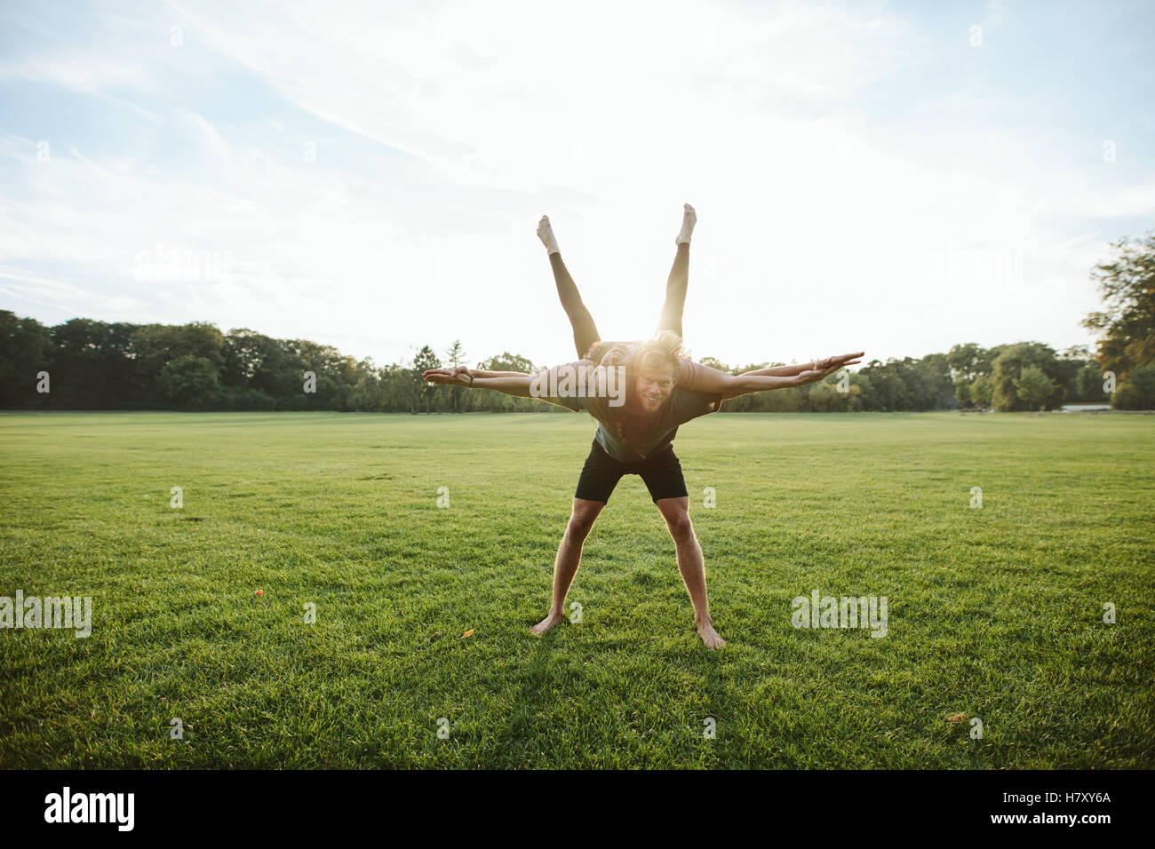 Healthy young couple doing acrobatic yoga workout in park. Man carrying and balancing woman on his back with their hands outstre Stock Photo