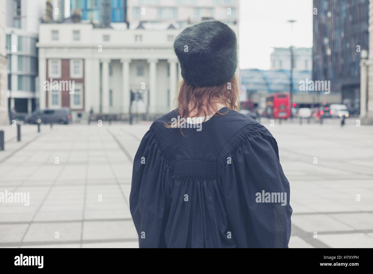 A young woman wearing a fur hat and a black graduation gown is standing in a square Stock Photo