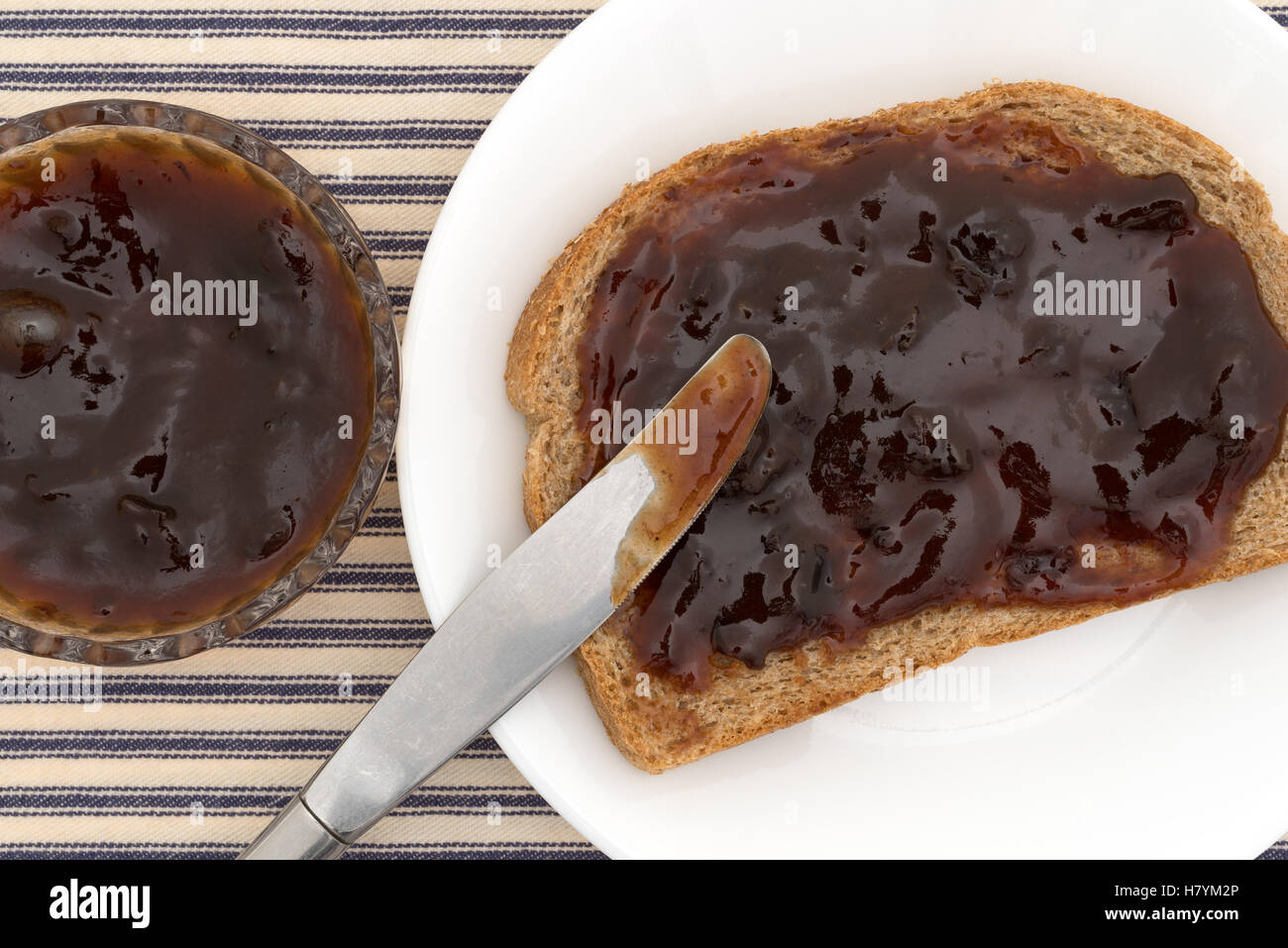 Top close view of plum preserves spread on a piece of wheat bread atop a white plate with a butter knife Stock Photo