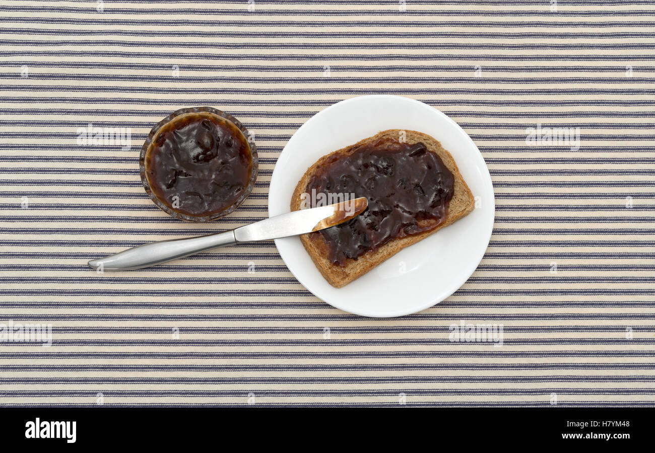 Top view of plum preserves spread on a piece of wheat bread atop a white plate with a butter knife and more jam to the side on a Stock Photo